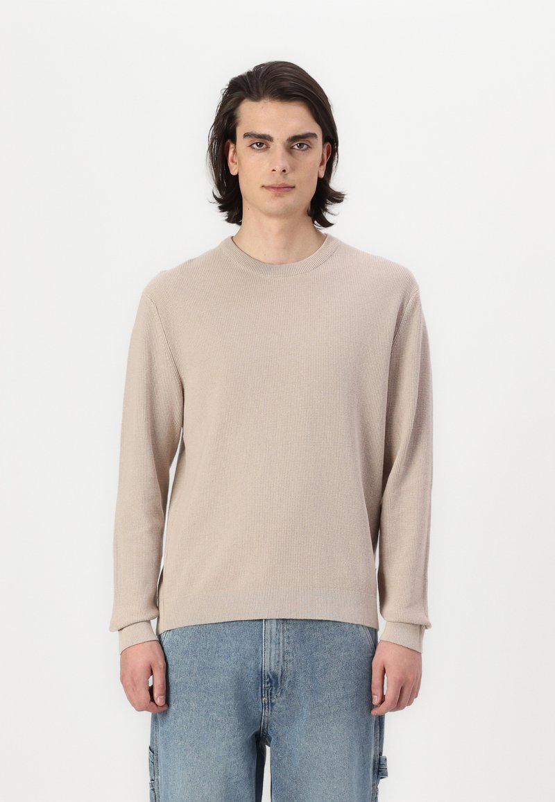 Maglione lungo beige a maniche lunghe con un design a maglia testurizzata, colletto rotondo e polsini. Abbinato a pantaloni in denim blu chiaro.