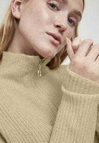 Beige hooggesloten trui met een gestructureerd, ribbelpatroon. Voorzien van een halve rits met een zilver kleurige ringtrekker. Close-up van de stof en handdetail.