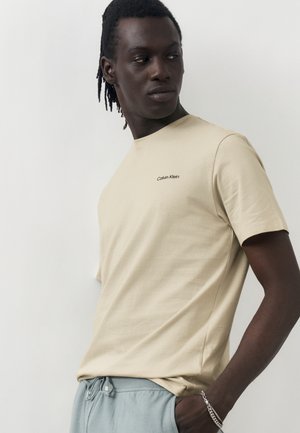 Mężczyzna w beżowej koszulce Calvin Klein i jasnoniebieskich spodniach, patrzący przez ramię z ręką w kieszeni na jednolitym tle.