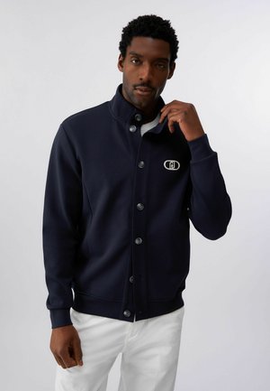 Uomo che indossa una giacca blu scuro abbottonata con un logo bianco sul petto e pantaloni bianchi, mentre sistema con una mano il colletto della giacca.