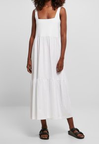 Sukienka maxi z białej bawełny, z kwadratowym dekoltem, bez rękawów oraz marszczoną, tiered spódnicą. Noszona z czarnymi sandałami.
