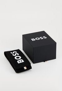 Scatola nera con la dicitura "BOSS" stampata in bianco. Sacchetto nero accanto ad essa, caratterizzato dallo stesso logo e da un piccolo etichetta con dettagli dorati.
