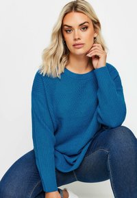 Blå stickad tröja med en lös passform och ribbad textur. Har en rund halsringning och långa ärmar. Kombineras med mörka jeans och vita sneakers.