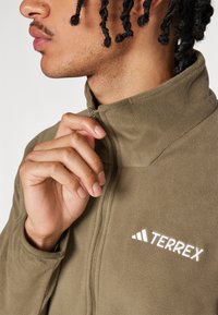 Zelená flísová bunda s vysokým golierom, s bielym logom "TERREX" a zipsom, zdôrazňujúca mäkkú textúru a priliehavý strih.
