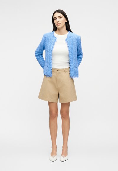 Γυναίκα που στέκεται με τα χέρια πίσω από την πλάτη, φορώντας ένα γαλάζιο υφασμάτινο σακάκι, λευκό τοπ, μπεζ σορτς και λευκές μυτερές γόβες.