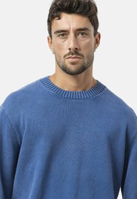 Man met kort krullend bruin haar en een baard, droeg een blauwe gebreide trui, kijkt recht vooruit met een neutrale uitdrukking en kleine ringoogsteker.