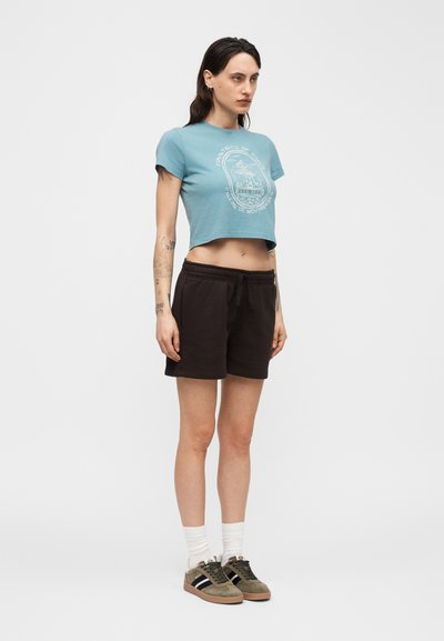 Γυναίκα όρθια, φορώντας μπλε κοντομάνικο μπλουζάκι με γραφικό σχέδιο, μαύρα σορτς με κορδόνι, λευκές κάλτσες και καφέ αθλητικά παπούτσια σε απλό λευκό φόντο.