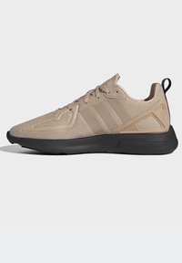 Beige athletic shoe with mesh upper, three tonal stripes, textured overlays, and a black sole. Features a pull tab and lace-up design.