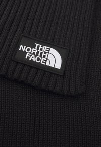 Bonnet en tricot noir à côtes présentant un patch rectangulaire noir avec du texte blanc et un logo, mettant en valeur une surface texturée et un design plié.