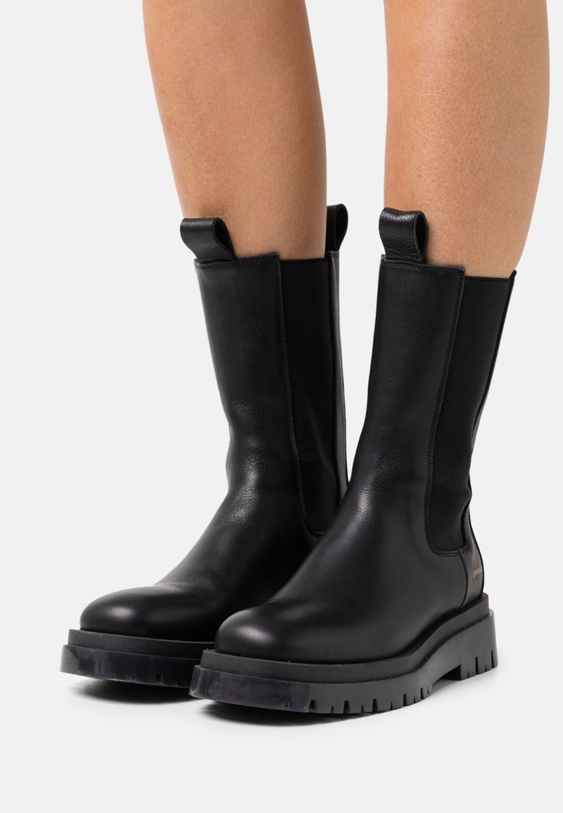 Bottes en cuir noires mi-mollet avec des semelles épaisses à crampons portées sur des jambes nues sur fond blanc.