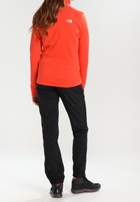 Chaqueta de forro polar naranja brillante con cuello alto, pantalones ajustados negros y zapatos de escalada negros con acentos morados. Perspectiva desde la parte trasera.