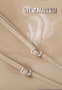 Borsa a mano in pelle verniciata beige con hardware argentato, dotata di due manici testurizzati e logo in rilievo. Superficie liscia e lucida.