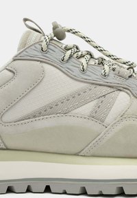 Zapatilla deportiva de color gris claro que combina materiales de malla y gamuza, con cordones estampados y un diseño de suela en capas con tracción moderada.