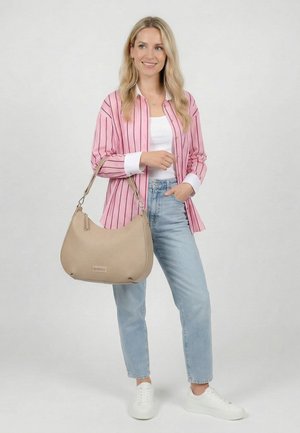 Hymyilevä nainen, jolla on päällään vaaleanpunaraidallinen paita ja farkut, pitää beigeä käsilaukkua olallaan, seisoo yksivärisen vaalean taustan edessä.