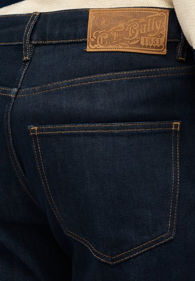 Εγγύς λήψη πίσω τσέπης από σκούρο μπλε τζιν με πορτοκαλί ραφές και καφέ δερμάτινη ετικέτα της μάρκας Bally στη μέση.