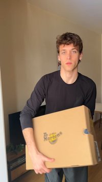 Caixa de cartão castanho com o logótipo "Dr. Martens", segurada por uma pessoa com uma camisa preta de mangas compridas. O fundo apresenta paredes e mobília em cores neutras.