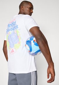 Hvid T-shirt med farverigt grafikdesign på ryggen, grå shorts og en blå fodbold med røde og orange accenter, holdt mod en arm.