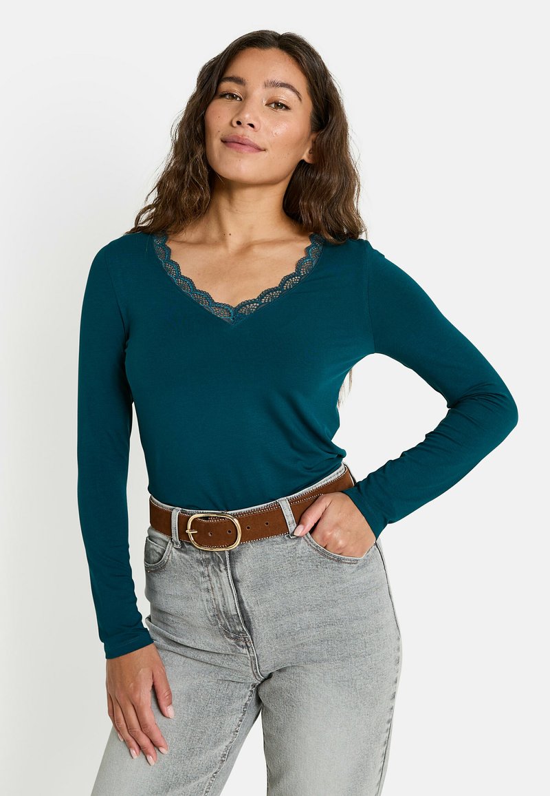 Camisa de manga larga en color verde azulado con escote de encaje con forma de ola, combinada con jeans gris claro y un cinturón marrón, presentando un corte clásico y una textura suave.