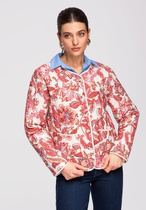 Kvinde iført en hvid quiltet jakke med røde blomsterprint over en blå skjorte med krave, poserer med hænderne nær jakke-enden mod en ensfarvet baggrund.