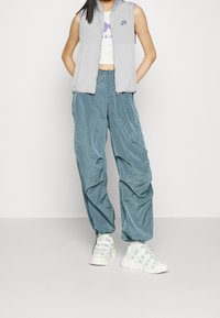 Ljusgrå ärmlös väst med blixtlås framtill, Nike-logotyp. Aqua textured baggy byxor. Vita klumpiga sneakers med mintgrön accent.