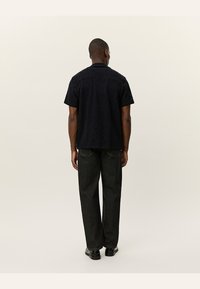 Camisa preta de manga curta com tecido texturizado. Usada com jeans escuros e folgados e sapatos pretos, vista por trás. Design de colarinho simples.