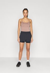 Débardeur marron assorti à un short de sport noir. Le modèle est debout, pieds nus, portant des baskets noires avec des chaussettes blanches. Design simple et ajusté.