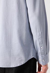 Poignet de chemise à manches longues bleu clair avec détails de boutons portée sur un pantalon noir, montrant l’avant-bras inférieur et le côté du torse.