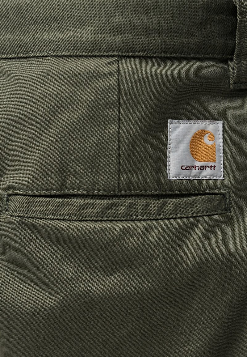 Pantalon en tissu vert olive doté d'une poche arrière avec une étiquette blanche affichant un logo jaune et le nom de la marque, cousue proprement.