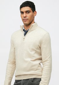 Cardigan beige zippé avec un col haut, fabriqué à partir d'un tissu doux, doté de manches longues et d'une coupe ajustée. Porté par-dessus une chemise à col.