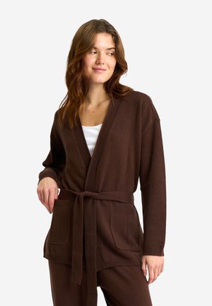 Kvinde med bølget brunt hår iført en brun cardigan med bælte over en hvid top, som ser til siden med et let smil og hånd i lommen.