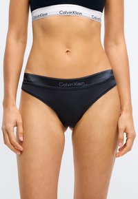 Schwarze Baumwoll-Bikini-Briefs mit einem satinähnlichen Bund, der das "Calvin Klein"-Logo zeigt. Glatte Textur, niedriger Sitz, minimalistisches Design mit wenig Bedeckung.