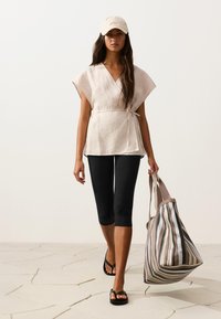 Femme portant un haut beige croisé, des leggings noirs courts, une casquette beige, des sandales noires, portant un grand sac fourre-tout rayé, marchant sur un sol carrelé clair.