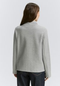 Grijze, textuurjasje met een hoge kraag en lange mouwen. Heeft een iets getailleerd ontwerp en een gladde afwerking aan de onderkant.