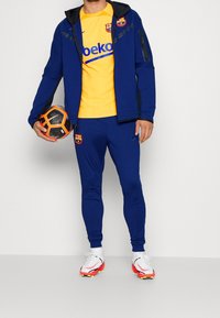 Tuta sportiva blu con giacca con zip e pantaloni lunghi, maglietta gialla con logo, scarpe chiodate nere e rosse, e tiene in mano un pallone da calcio nero e arancione.