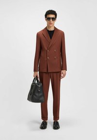 Braunes doppelt geknöpftes Blazer, kombiniert mit passenden Hosen. Schwarzes Hemd darunter, hält eine strukturierte schwarze Tasche und trägt Sonnenbrillen.