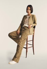 Veste bomber olive clair, t-shirt blanc et pantalon kaki à motifs avec découpes ; assis sur une chaise en bois, des sneakers blanches complètent le look.
