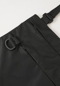 Svart nylonveske med to glidelåslommer, en teksturert overflate og justerbar stropp med plastdetaljer og løkke.