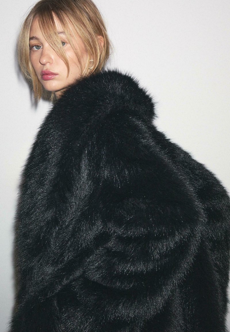 Crni kaput od umjetnog krzna s debelom, mekanom teksturom. Kaput ima zaobljen ovratnik i voluminozan, oversized kroj.