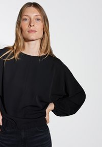 Blusa nera con scollo rotondo, maniche a palloncino e vita cintrata. Tessuto morbido con un leggero lucido, abbinata a jeans scuri.
