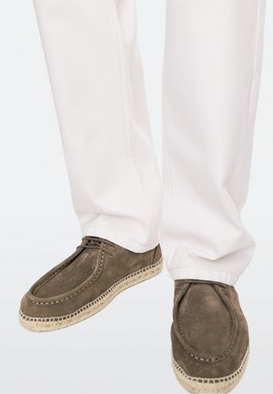Braune Wildleder-Espadrilles mit gesteppten Details, getragen mit weißen Hosen vor neutralem Hintergrund.