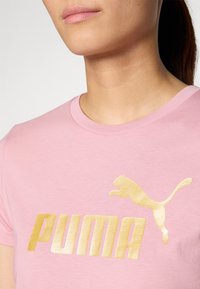 Rožinė medvilninė marškinėlė su auksiniu PUMA logotipu ir šokančiu katinu. Marškiniai turi apvalų apykaklę ir lygią tekstūrą.