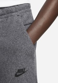 Šedé tepláky Nike s texturovanou fleecovou látkou, vybavené boční kapsou a černým logem Nike na levém stehně.