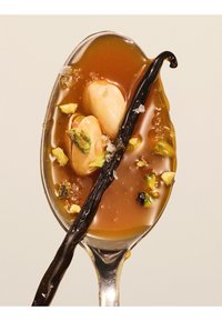 Cuillère avec sauce caramel, deux amandes, pistaches hachées, sel grossier et une gousse de vanille noire sur un fond clair.