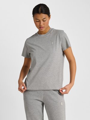 Woman adjusting hem of plain grey short-sleeve t-shirt, wearing matching grey pants, against plain light background.