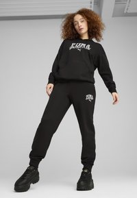 Zwarte hoodie en sweatpants set gemaakt van zachte stof, met een opvallend wit Puma-logo op de borst en zijkant. Stevige zwarte sneakers.