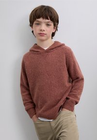 Felpa con cappuccio in maglia color ruggine con finitura testurizzata, polsini a costine e tasca a marsupio. Indossata sopra una camicia bianca e abbinata a pantaloni beige.