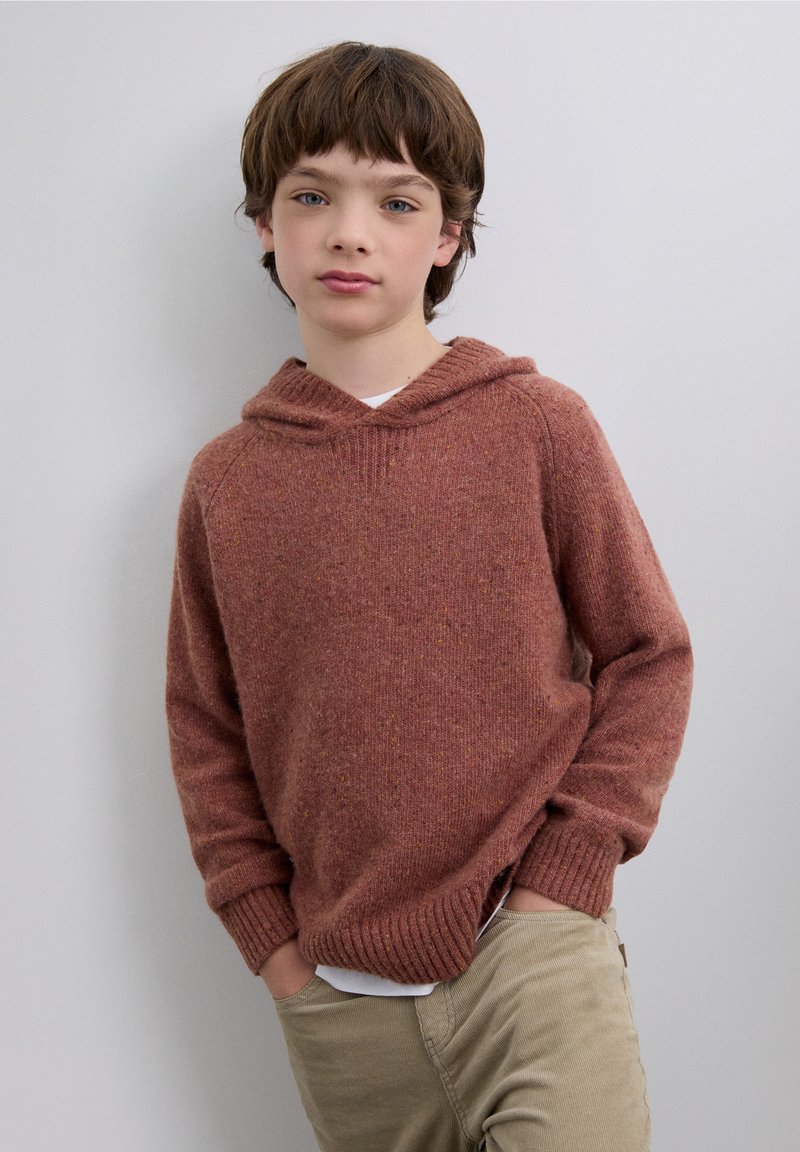 Felpa con cappuccio in maglia color ruggine con finitura testurizzata, polsini a costine e tasca a marsupio. Indossata sopra una camicia bianca e abbinata a pantaloni beige.