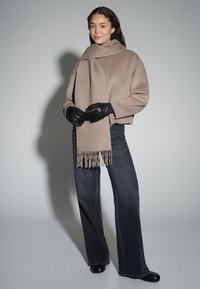 Beige Wollmischgewebe-Wickel mit Fransen, getragen mit schwarzen Lederhandschuhen und dunkelgrauen Jeans mit weitem Bein. Strukturierter Stoff, klare Linien und neutrale Töne.