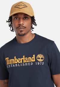 Námorně modré bavlněné tričko s žlutě vyšívaným textem a logem "Timberland". Khaki kordový klobouk s černým logem vpředu.