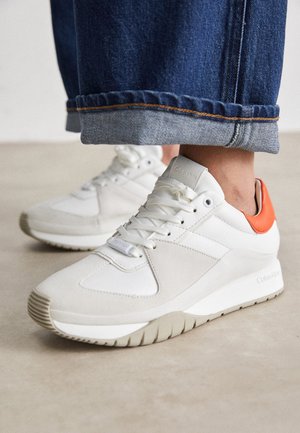 Zapatillas blancas y beige con detalles naranjas en el talón, usadas con jeans azules remangados sobre un fondo de suelo claro.